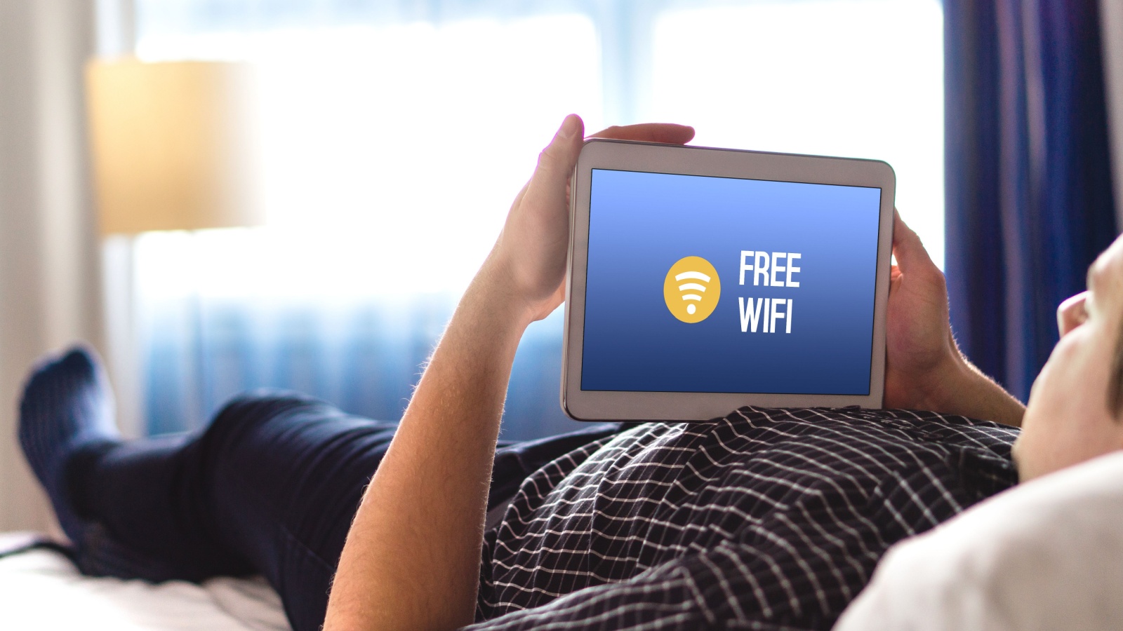 Man using free hotel wifi with tablet. Lying in hotel room bed and browsing internet. Public access online and connection available for customers,