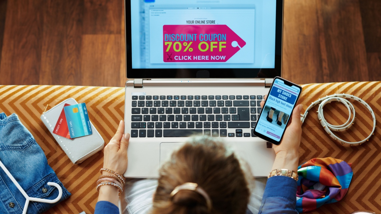 Upper view of young housewife in blue blouse browsing shoe retail online store on a smartphone and got email with discount coupon on a laptop while sitting on couch in the modern living room.