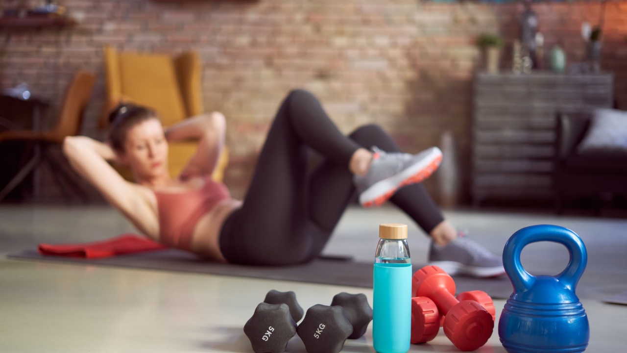 Focus on fitness equipments, barbell and kettlebell. Woman doing sit-ups in the background. Concepts about home workout, fitness, sport and health.