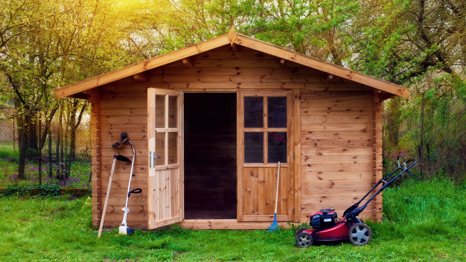 Garden shed (front view) with hoe, string trimmer, rake and grass-cutter. Gardening tools shed. Garden house on lawn in the sunset. Wooden tool-shed.