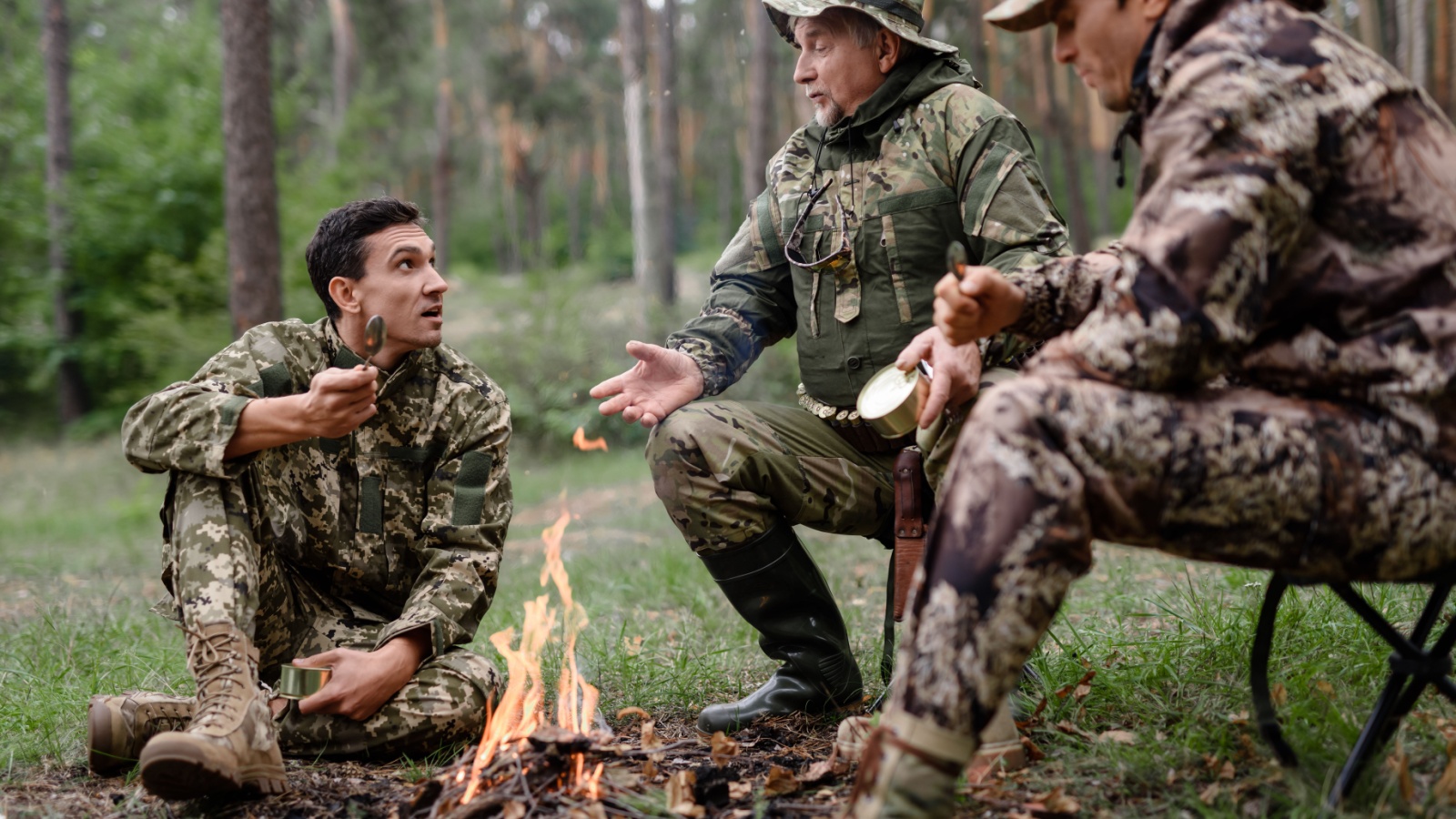 Hunters sit on a halt and discuss something behind a fire. Hunters are sitting near a campfire in camouflage uniforms.