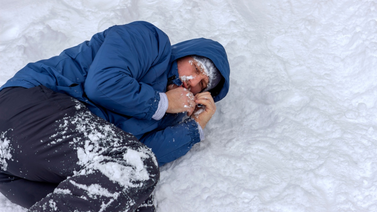 Frozen man in a blue jacket and hat lying down covered snow and frost, trying to stay warm on a very cold winter day, snow falls around him. Sick mountaineer with hypothermia on snow during the day. climate change