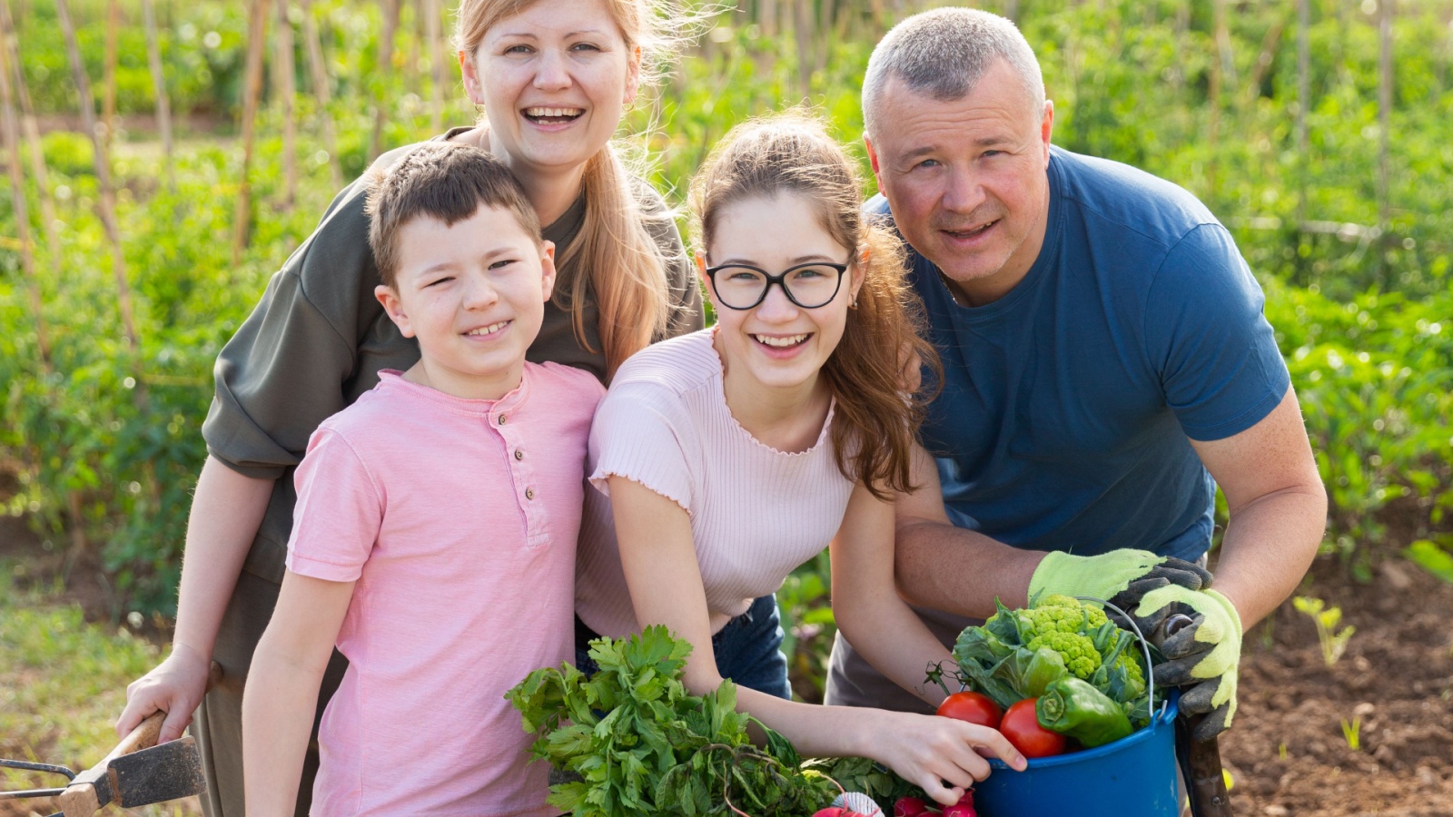 Portrait of happy family with two kids posing in garden with fresh harvest of vegetables and greens