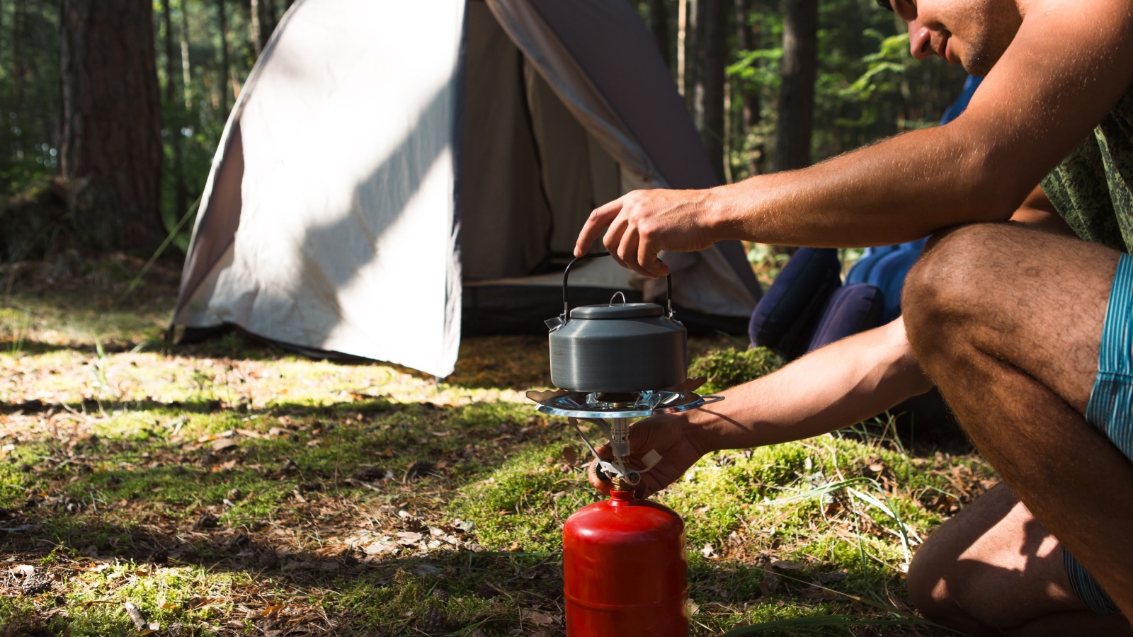 Cooking, heating kettle on a portable gas burner with a red gas cylinder. Camping, a man cooks breakfast outdoors. Summer outdoor activities