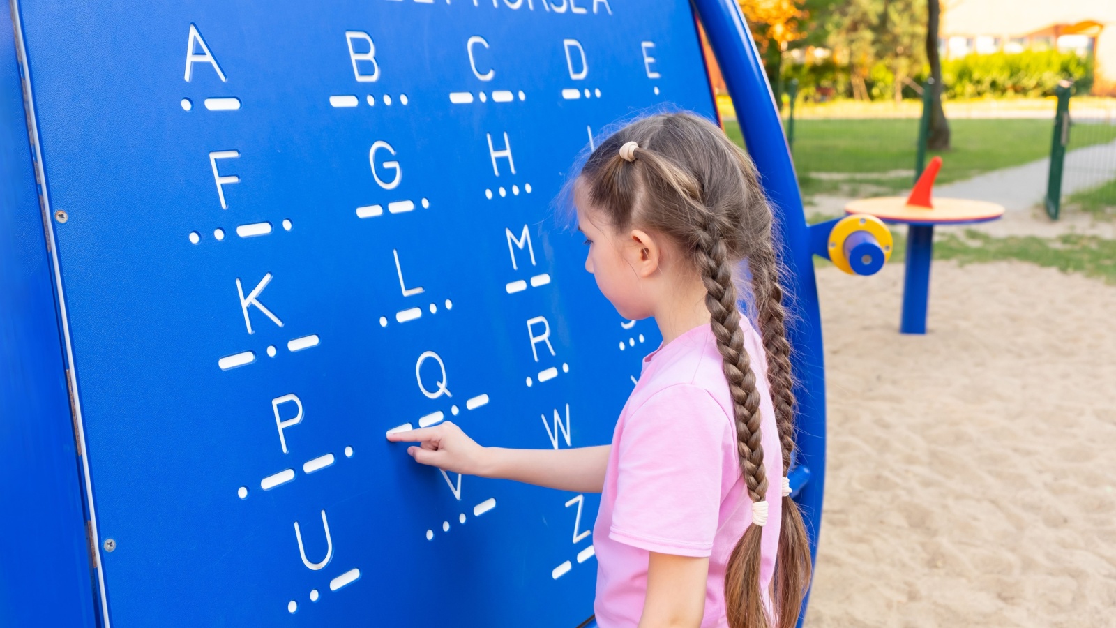 A little girl 7-8 years old with long hair dressed in a pink T-shirt and shorts is studying Morse code on a blackboard in the playground.