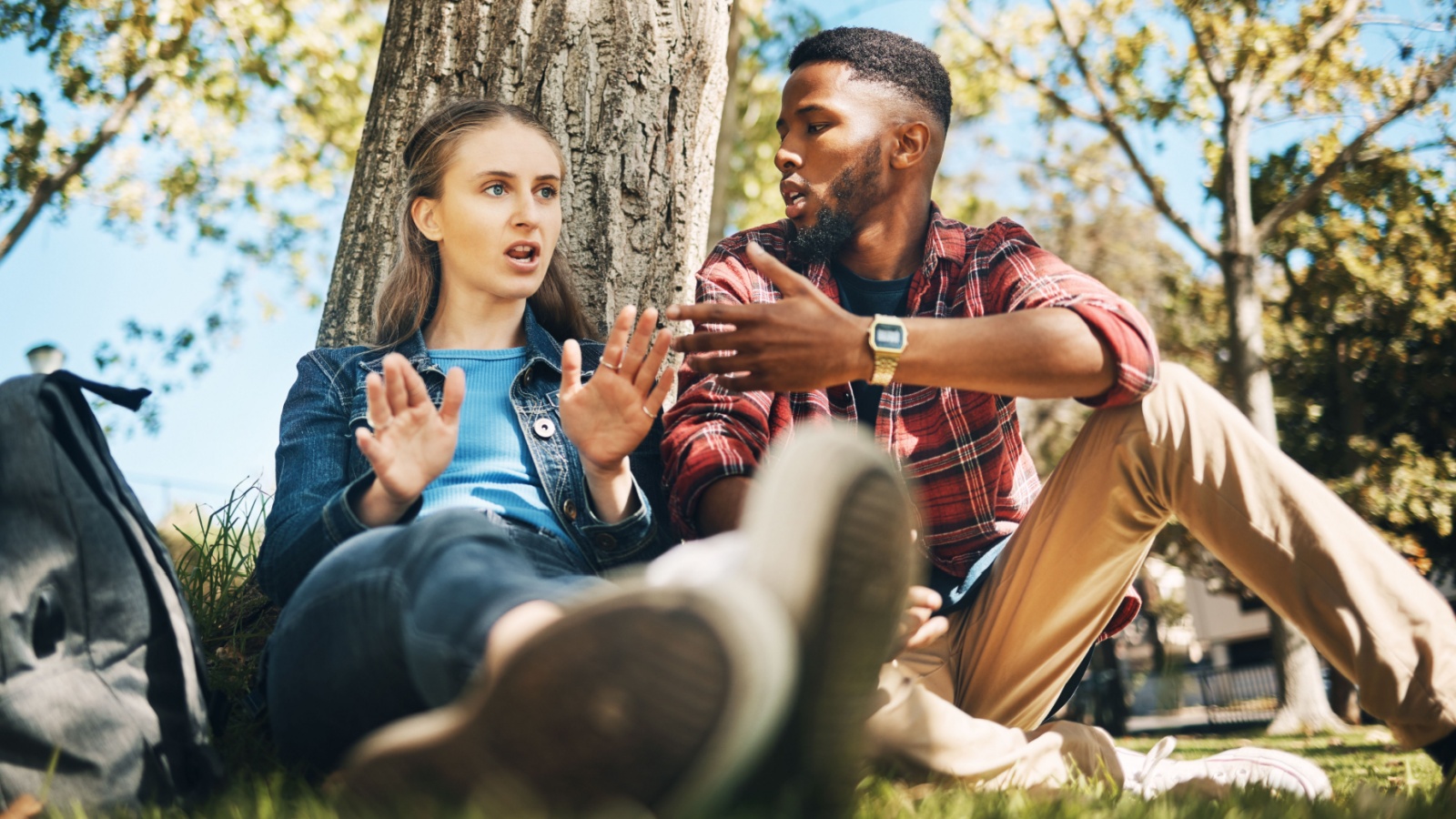 Interracial couple, fight and outdoor with upset young people angry on summer holiday. Conversation, conflict and break up discussion of a woman and man sitting by tree together talking about problem