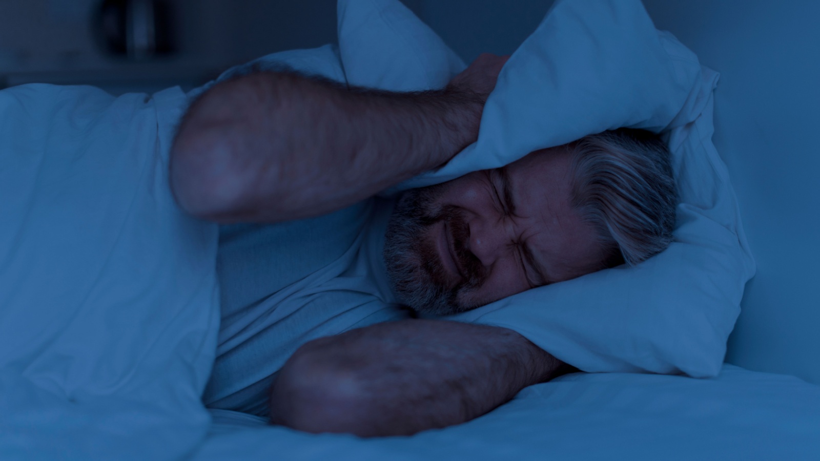 angry mature man lying in bed and covering head ears with pillow at night, cannot sleep, suffering from noisy neighbours or snoring spouse partner