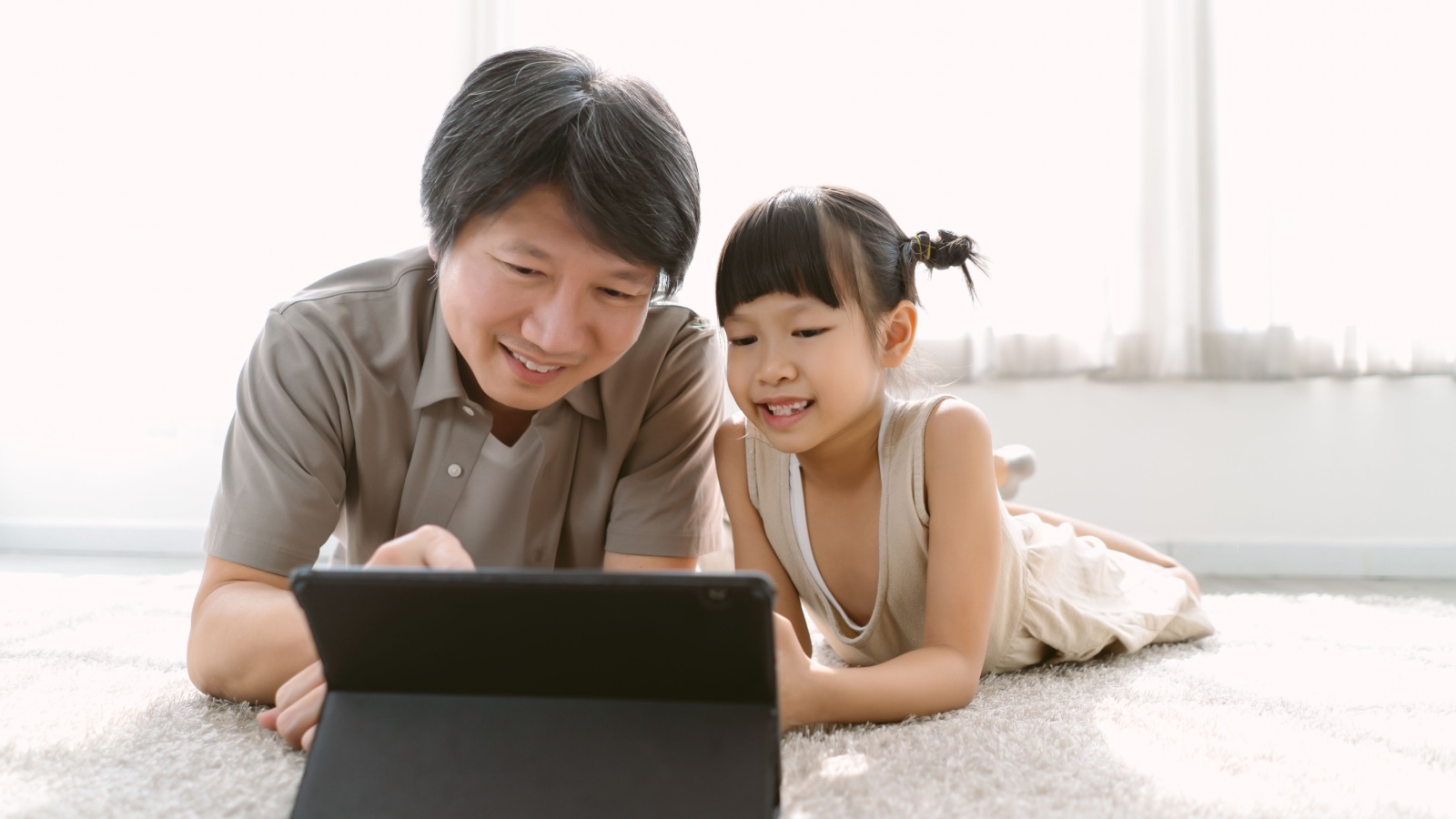 father and cute little daughter watching cartoons using digital tablet gadget e-books for kids on-line lying on carpet at home. Parental control protect from bad content