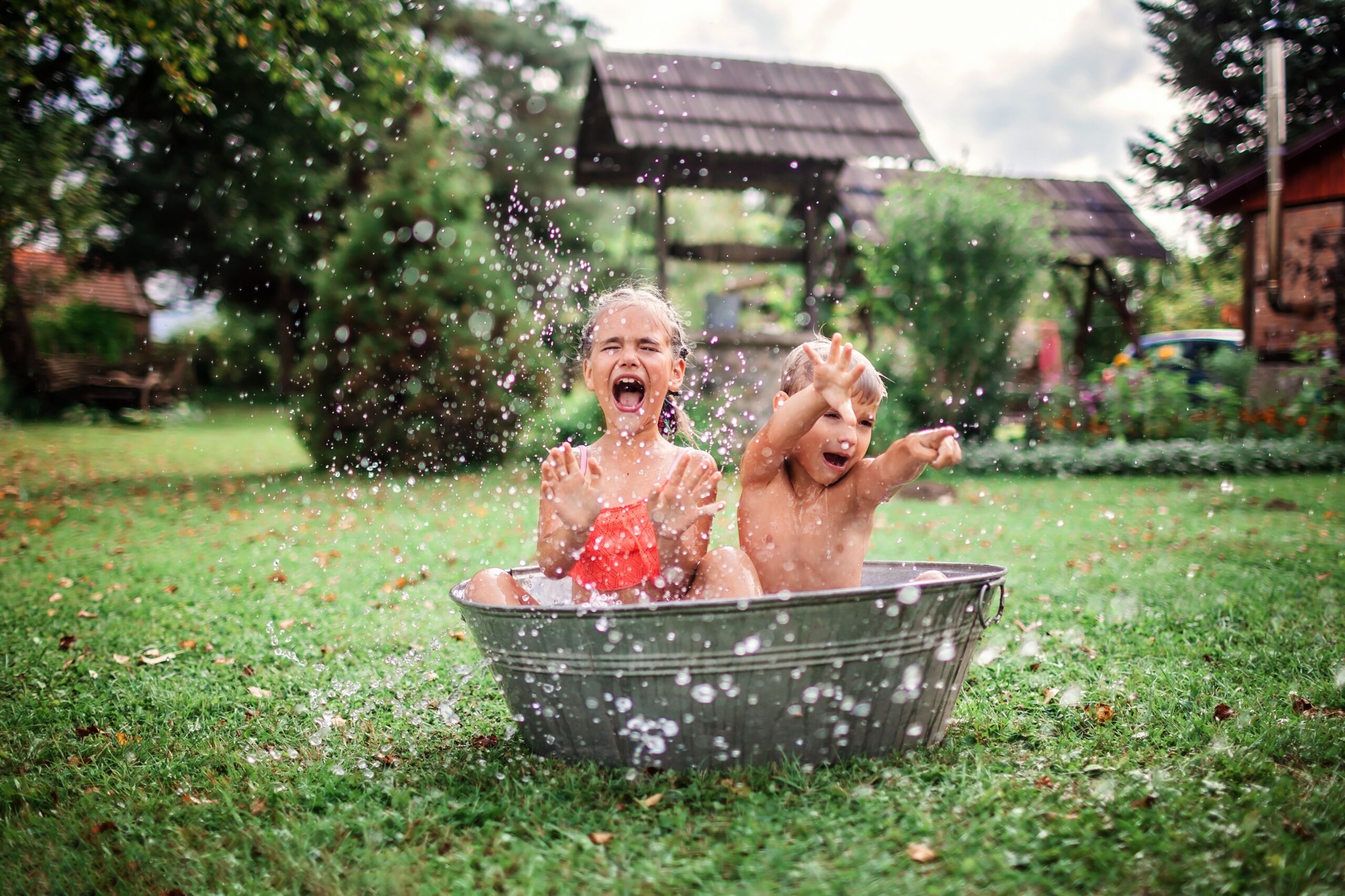 Cute kids having fun, bathing and splashing in old iron bowl on the backyard of wooden house, happy summertime, outdoor lifestyle, 