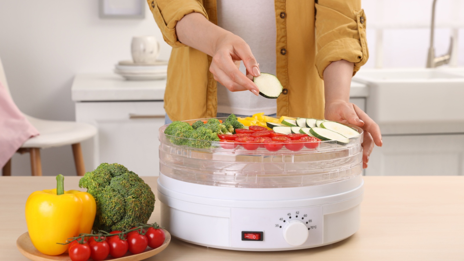 Woman putting cut zucchini into fruit dehydrator machine at wooden table in kitchen