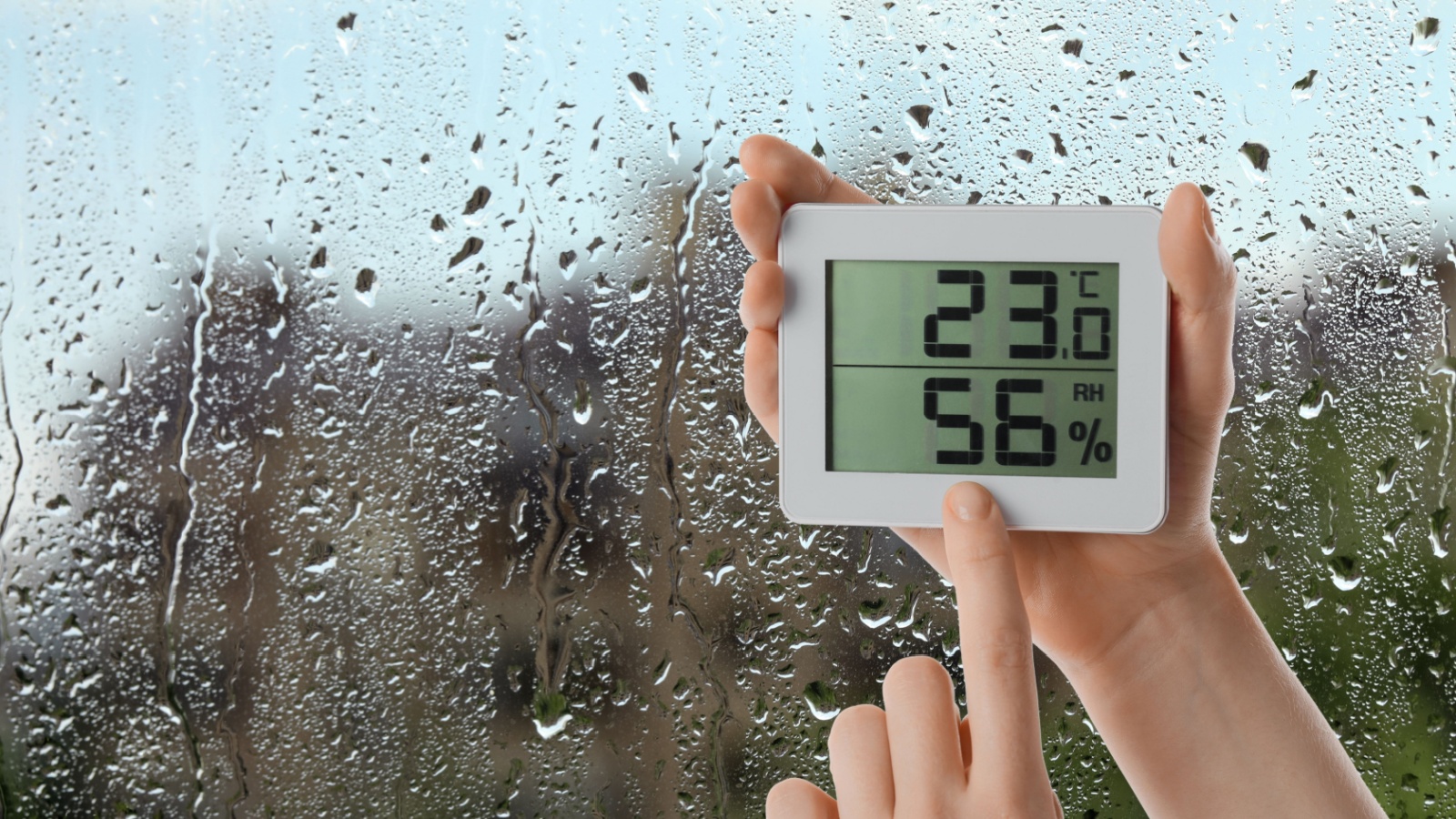 Woman holding digital hygrometer with thermometer near window on rainy day, 