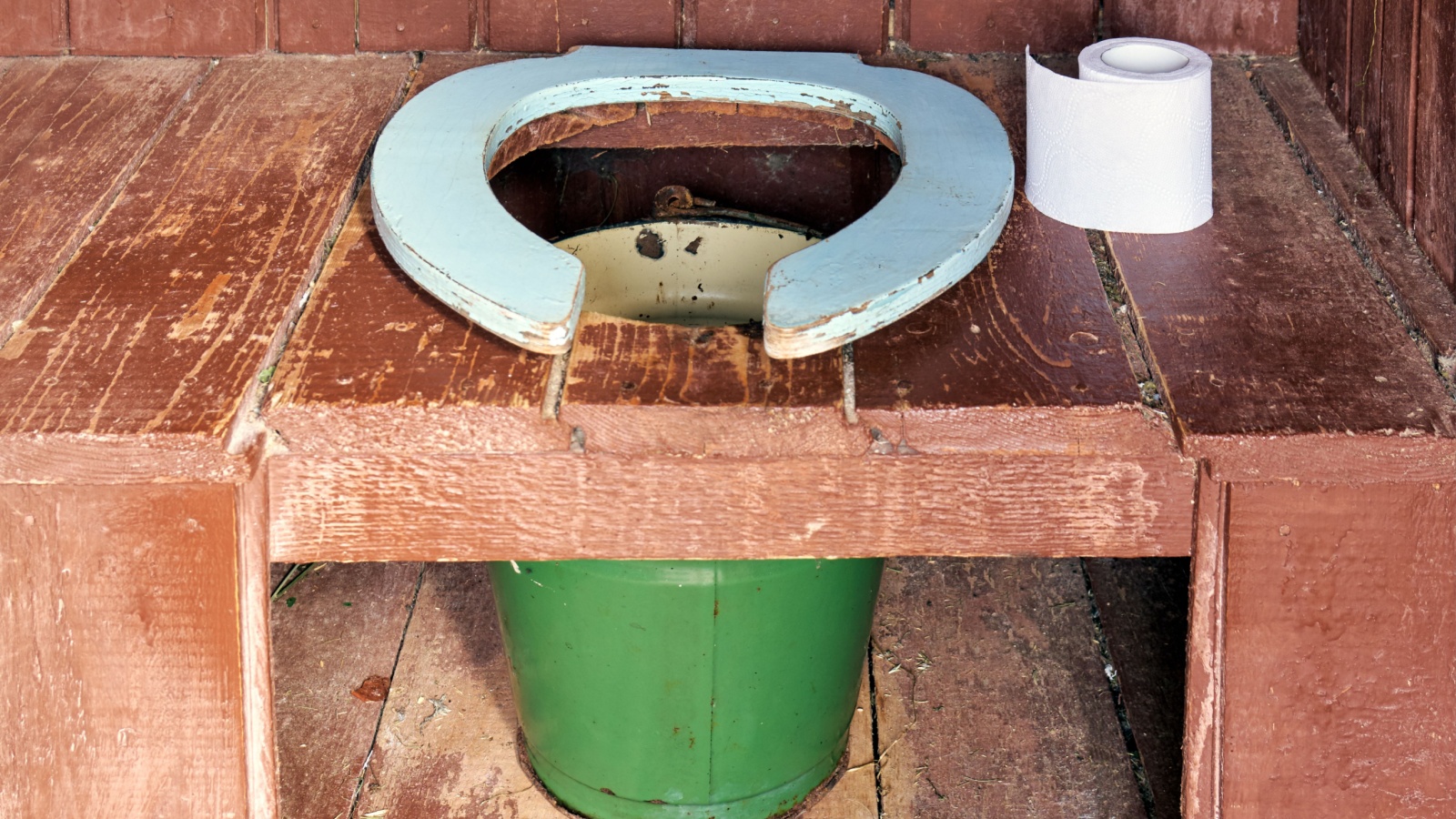 Toilet seat of a country wooden outdoor toilet with a metal bucket as a waste tank and a roll of a paper