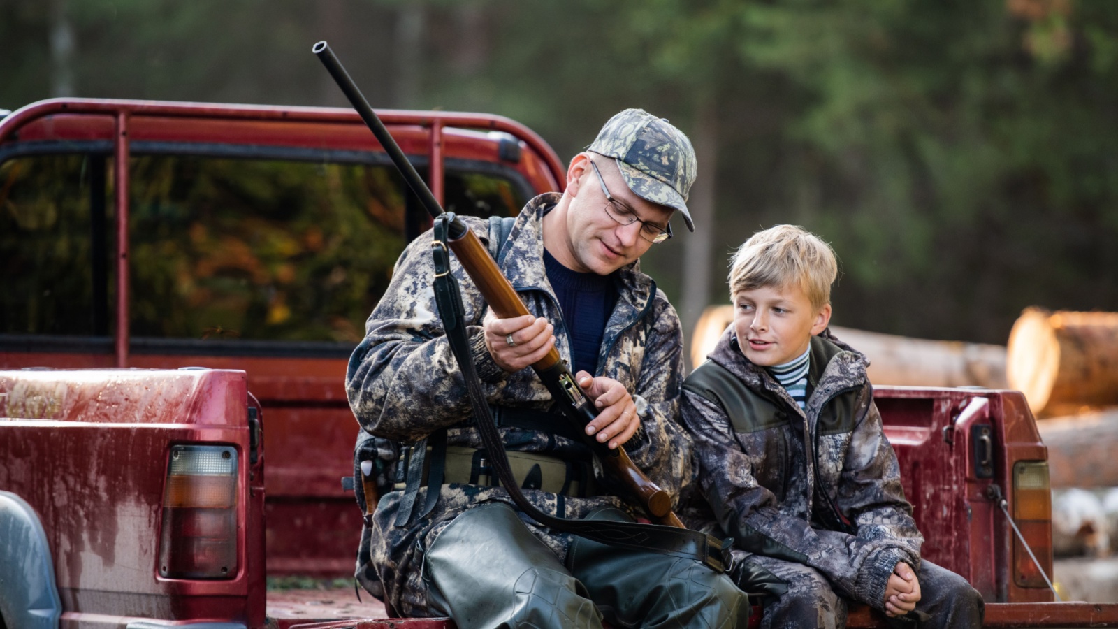 Father and son sitting in a pickup truck after hunting in forest. Dad showing boy mechanism of a shotgun rifle.