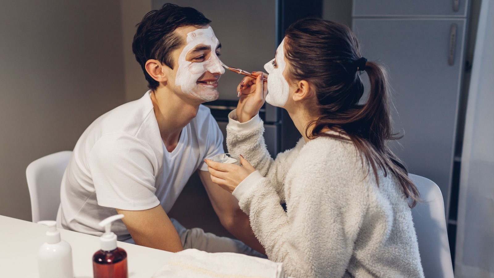 Woman applying clay mask on her husband's face. Young loving couple taking care of skin at home. Healthy routine