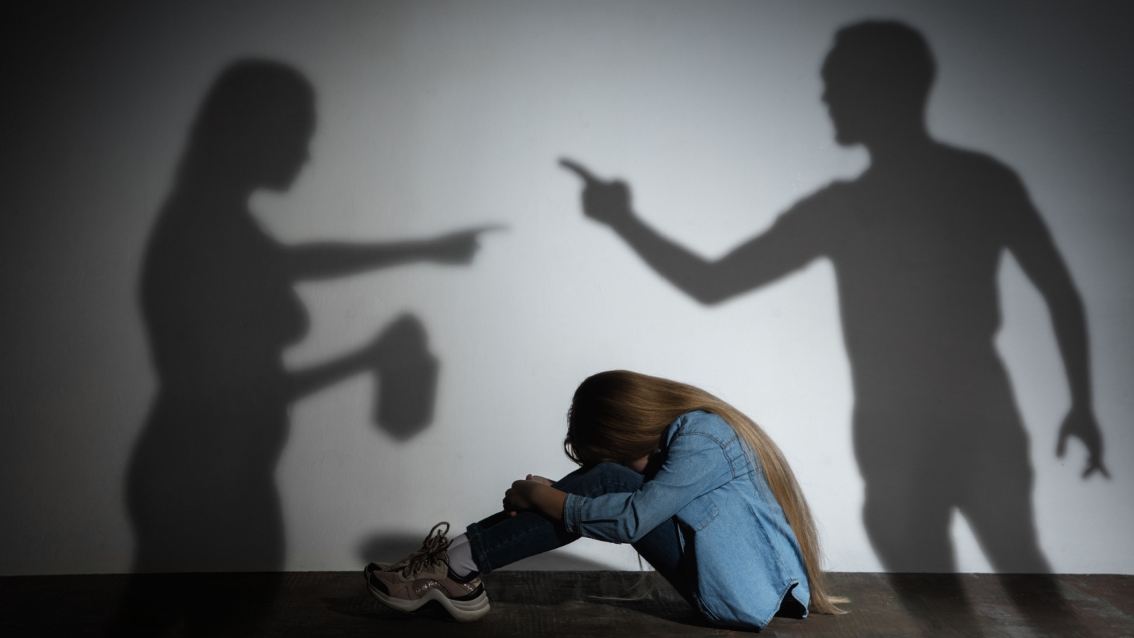 Domestic physical violence, abusing. Scared little caucasian girl, victim sitting close to white wall with shadow of angry threatening parents with alcohol addiction. Awareness of social problem.