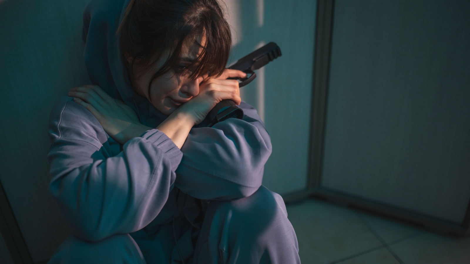 Upset woman sitting on the floor in the dark, holding a gun and crying after committing murder in self-defense; scared woman victim of domestic violence holding a gun for self-defense