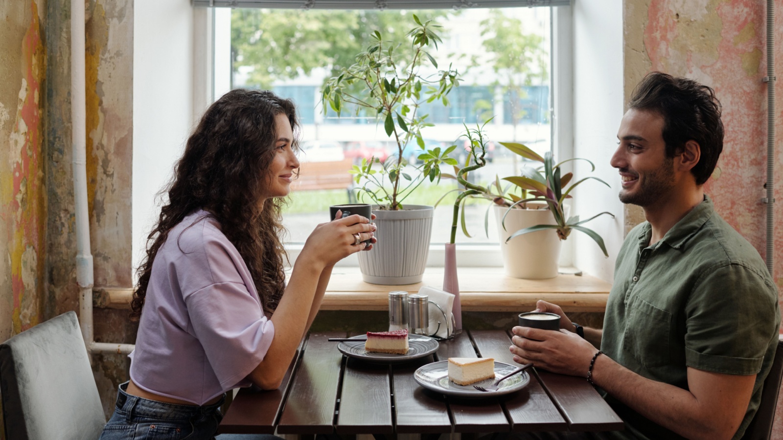 Young woman and her smiling boyfriend with cups of coffee sitting by table in front of one another and chatting