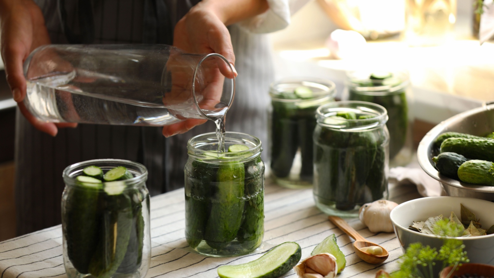 Woman pouring water into jar with canning cucumbers in kitchen,