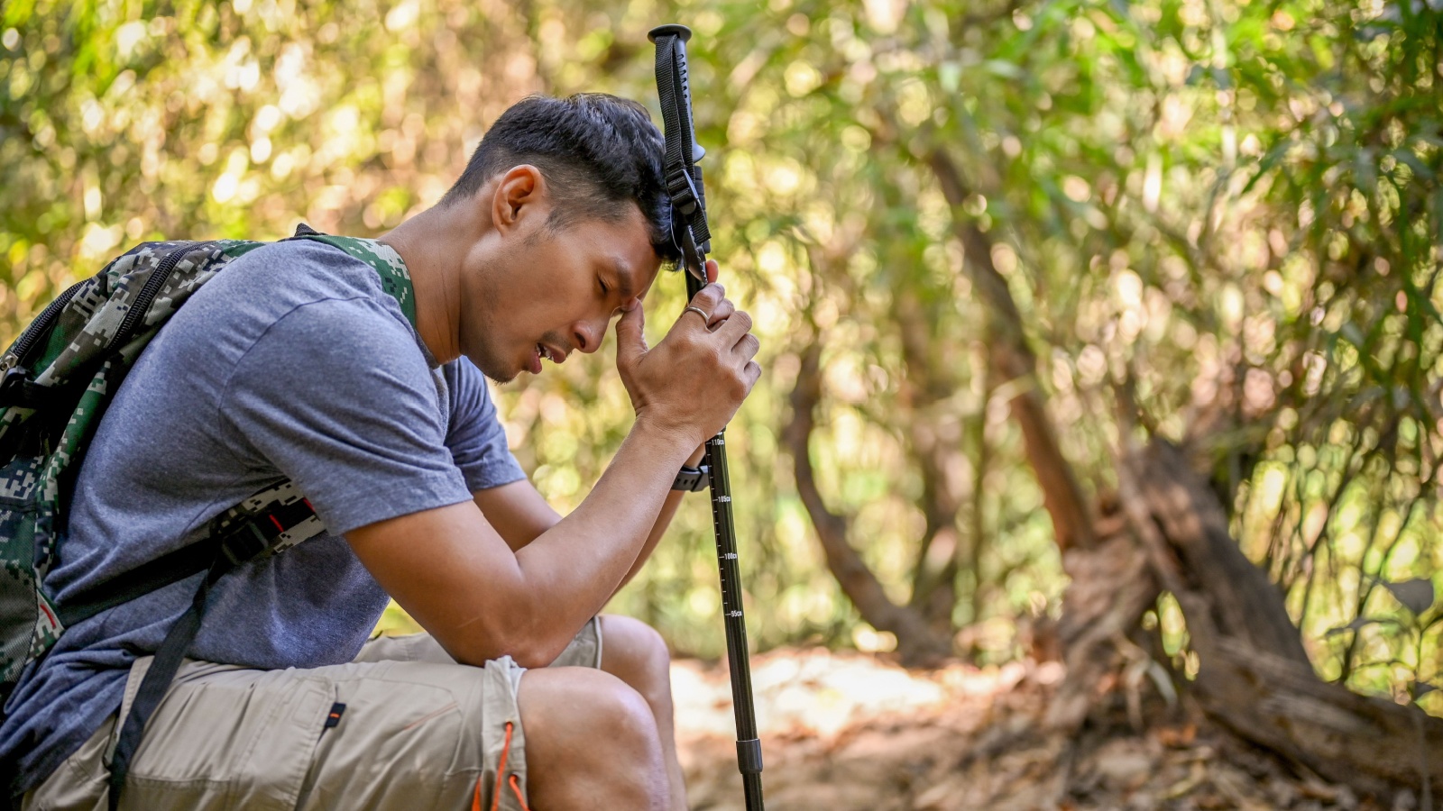 Tired and exhausted Asian male traveler with trekking gear sits on wooden log, resting while trekking in the forest.