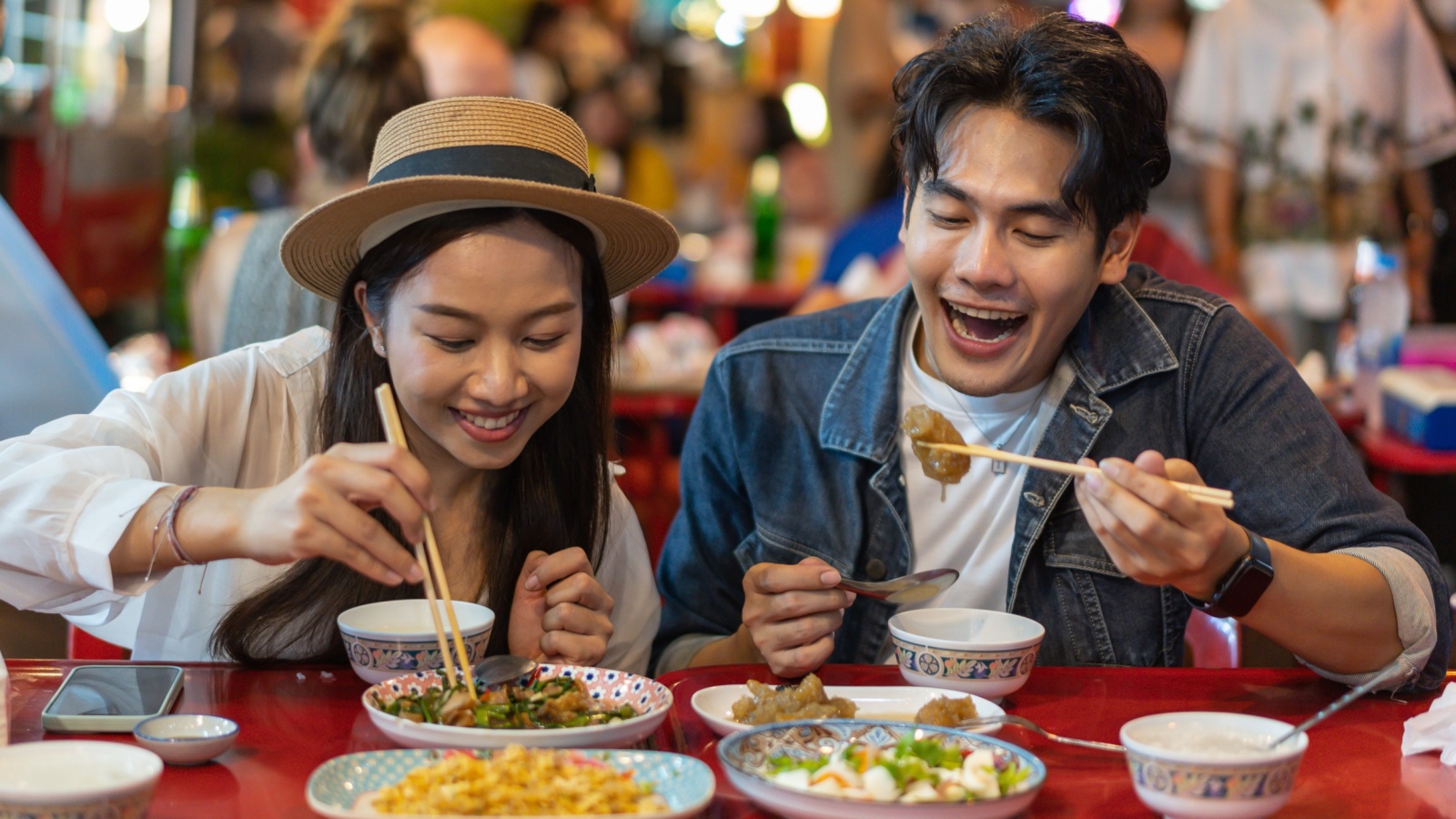 Young Asian couple traveler tourists eating Thai street food together in China town night market in Bangkok in Thailand - people traveling enjoying food culture concept