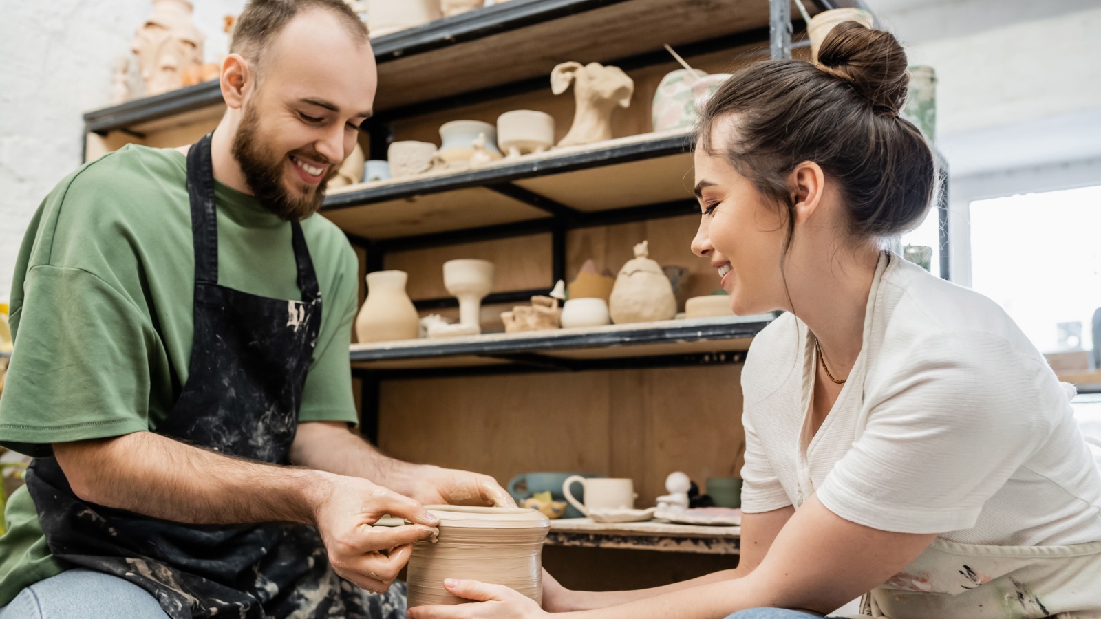 Smiling romantic artisans in aprons shaping clay vase together on pottery wheel in ceramic workshop