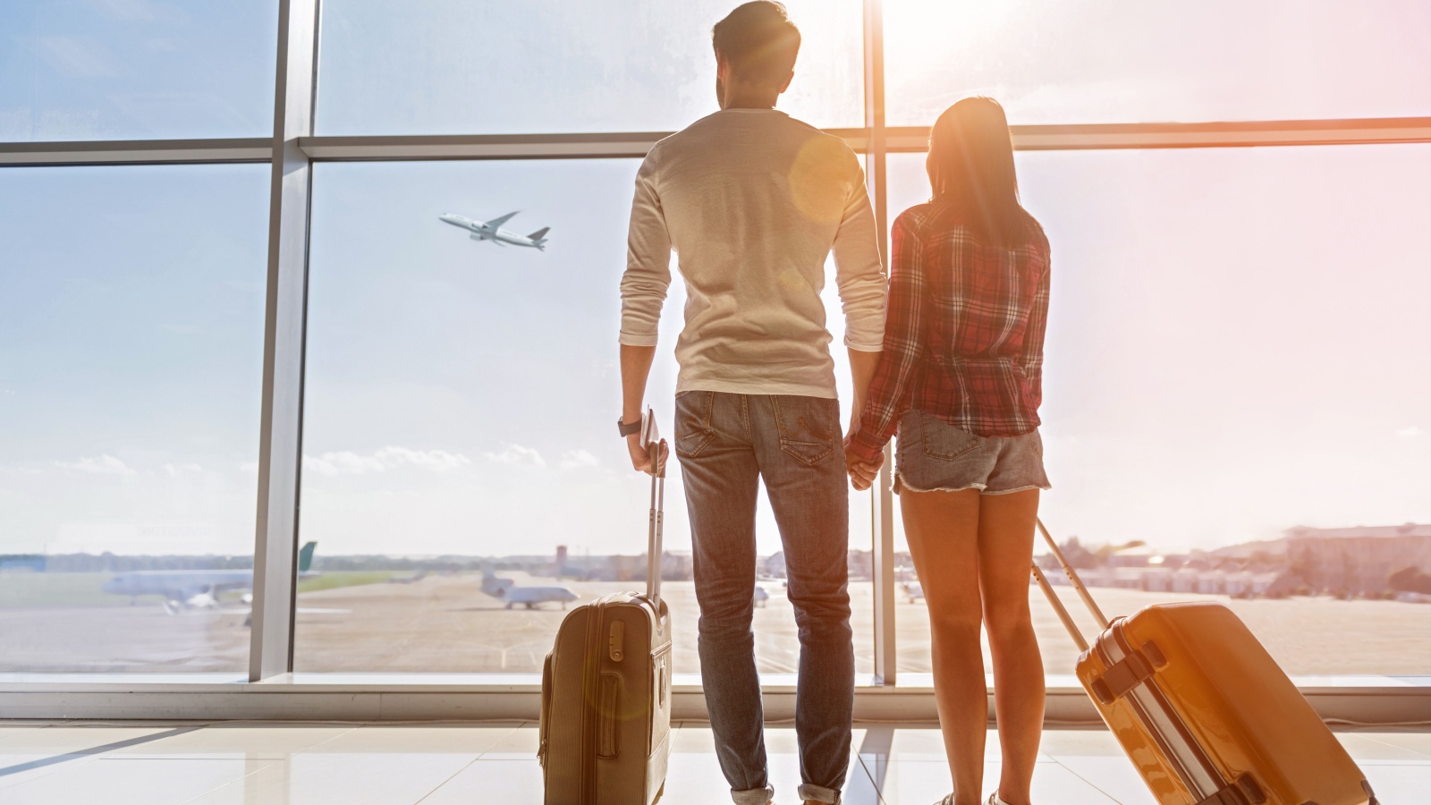 Inspired young loving couple is looking at flying plain in sky. They are standing near window at airport and holding hands