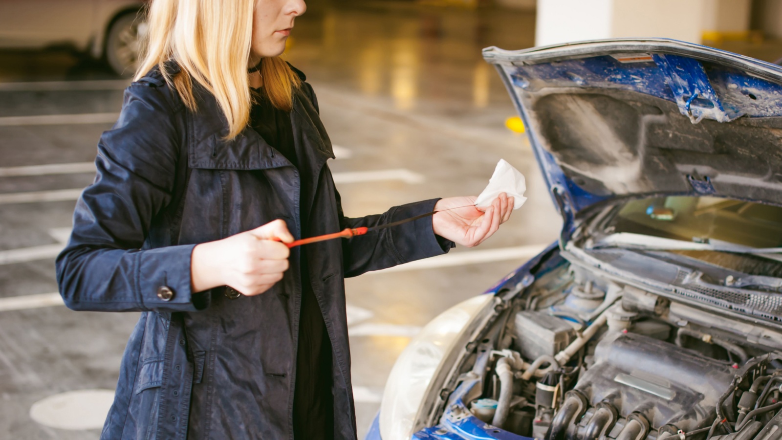 woman stands near car with raised engine compartment hood, checks engine oil level in engine, inspects feeler gauge